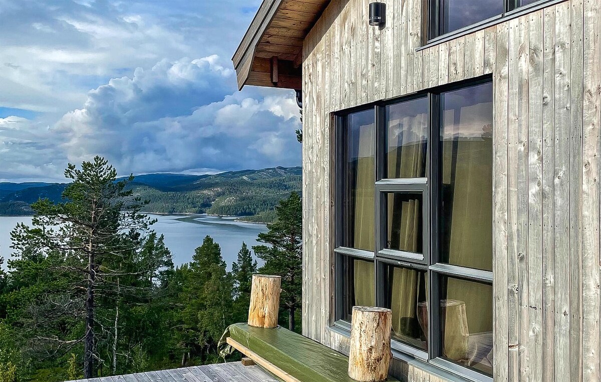 A wooden terrace showcases a green bench made from natural materials, offering a place to relax. Large windows reflect the surrounding landscape, revealing views of trees and a serene lake in the distance. Clouds drift softly across the sky, enhancing the calm ambiance.