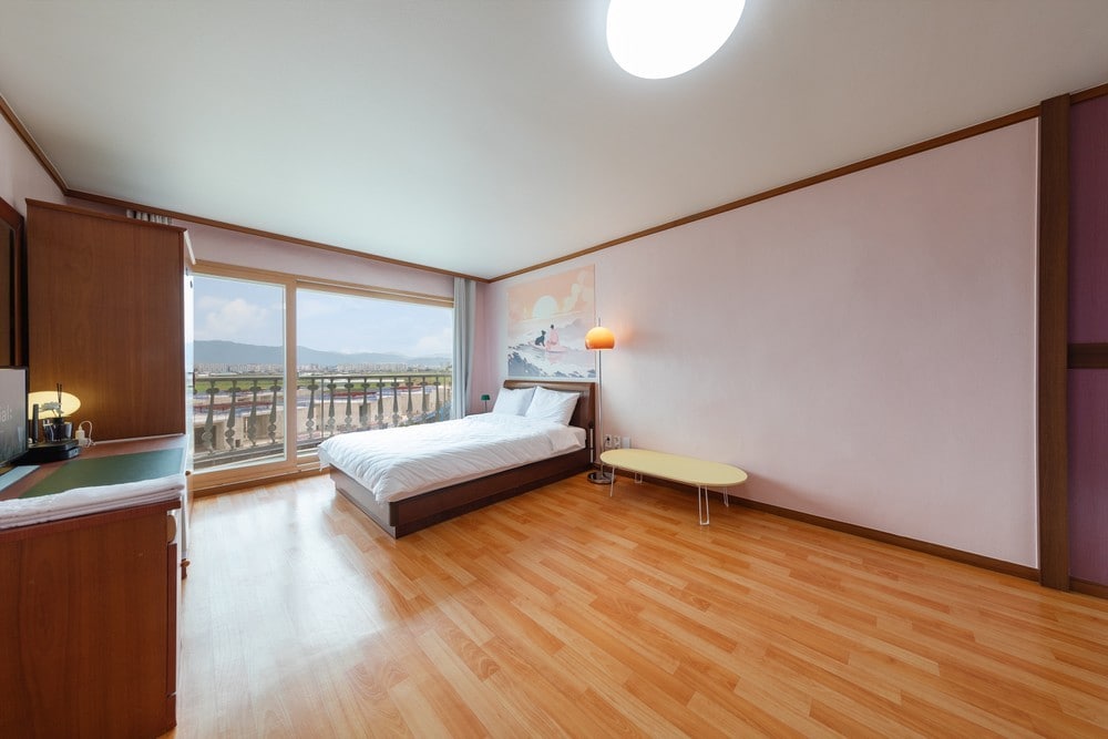 A spacious room features warm wooden flooring and light walls. A large window offers views of the outdoors, complemented by a comfortable bed and a small table. A subtle glow from a lamp enhances the tranquil ambiance without overwhelming the space.