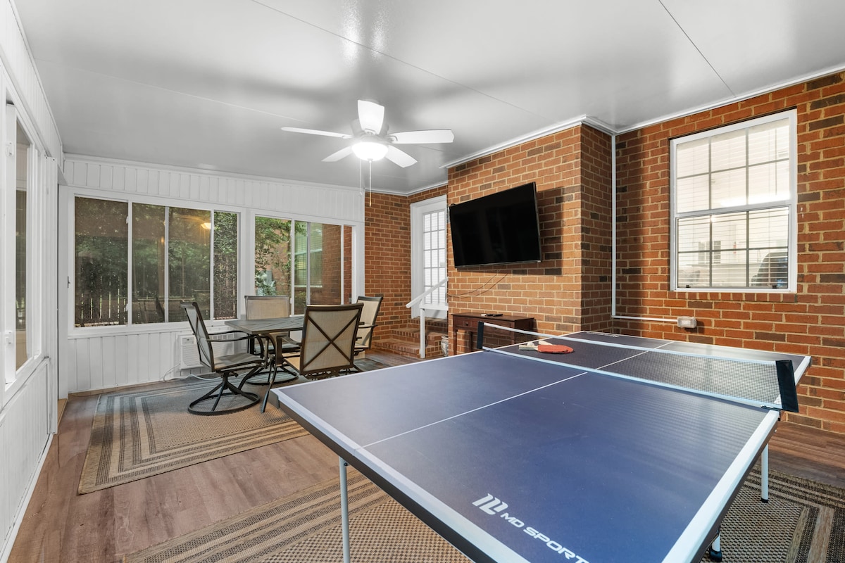 A screened-in patio area features a ping pong table, providing a space for recreational activities. Two windows invite natural light, while a ceiling fan offers airflow. A wall-mounted television and a dining set are also visible, enhancing the space for relaxation and entertainment.