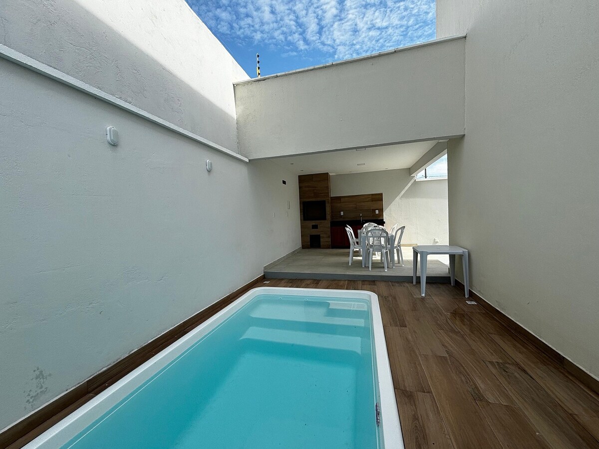 A brightly lit area features a small private pool surrounded by wooden flooring. A dining table with chairs is present on a raised platform, enhancing the space. The smooth walls are contrasted by a partially open ceiling, allowing natural light to fill the area.