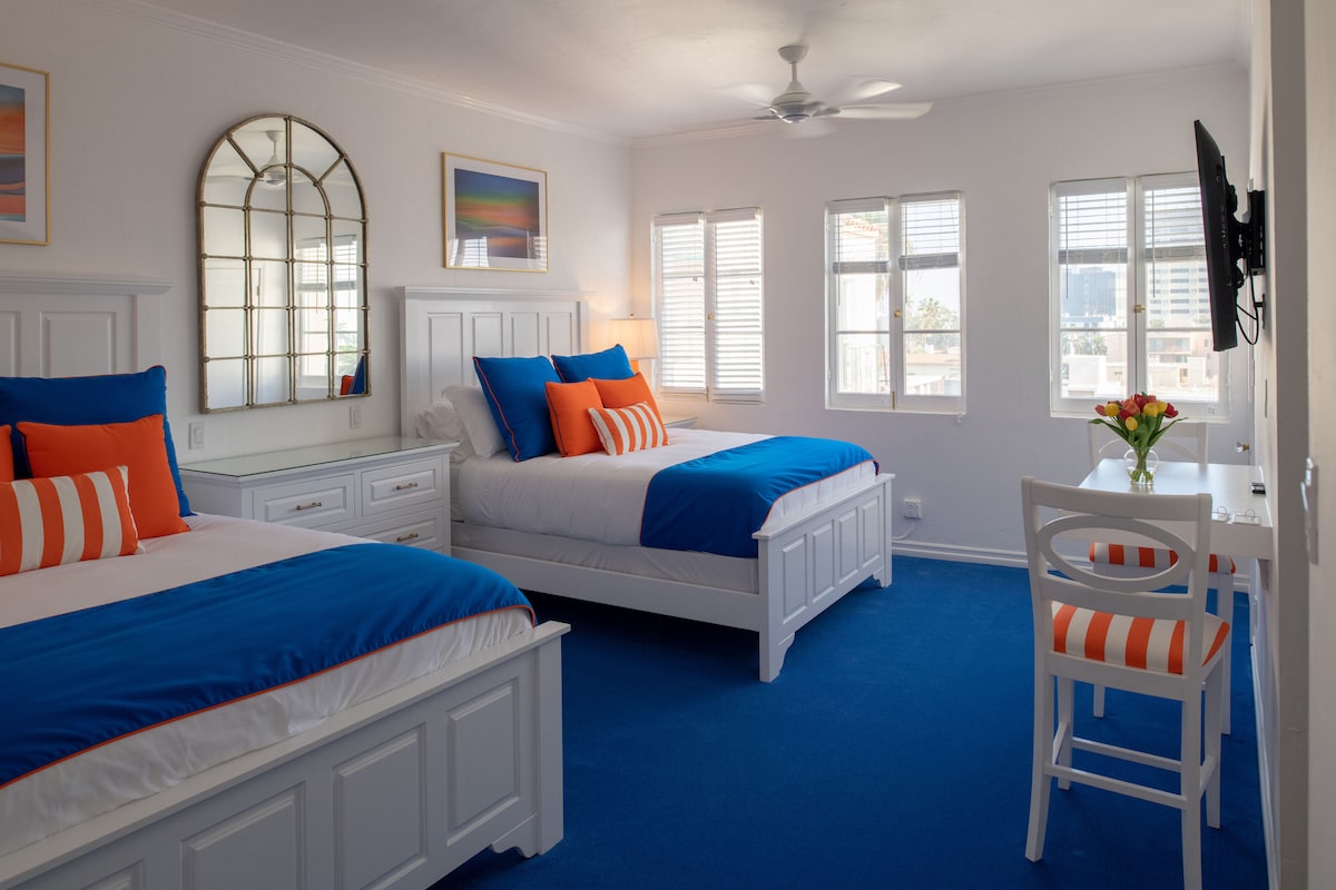 A bright room features two spacious queen beds dressed in blue and white linens, accented with orange pillows. A circular mirror reflects light from the windows, and a small table with a chair displays a flower arrangement. Natural light fills the space, highlighting the vibrant decor.