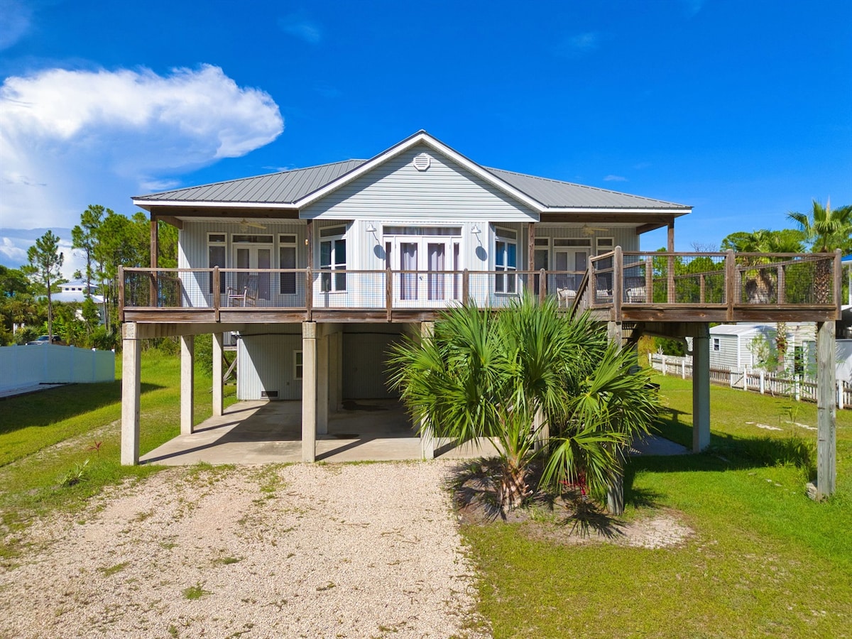 A raised home features a light blue exterior with a modern design. Multiple balconies are visible, supported by concrete pillars. Lush green grass surrounds the property, with palm trees adding a tropical touch. The blue sky is clear, enhancing the sense of openness.