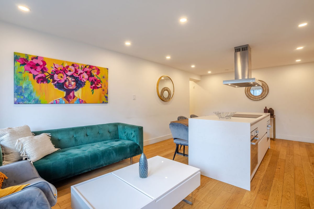 A stylish living area features a vibrant piece of artwork depicting a figure with flowers, set against a bright background. Comfortable seating options are arranged around a modern, minimalistic coffee table. A sleek kitchen island with a stovetop is visible in the background.