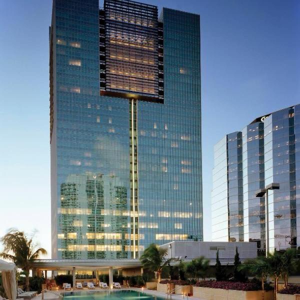 A modern high-rise building is shown, featuring a sleek glass façade that reflects the surrounding skyline. The pool area is visible in the foreground, framed by palm trees and lounge chairs, creating an inviting space for relaxation.