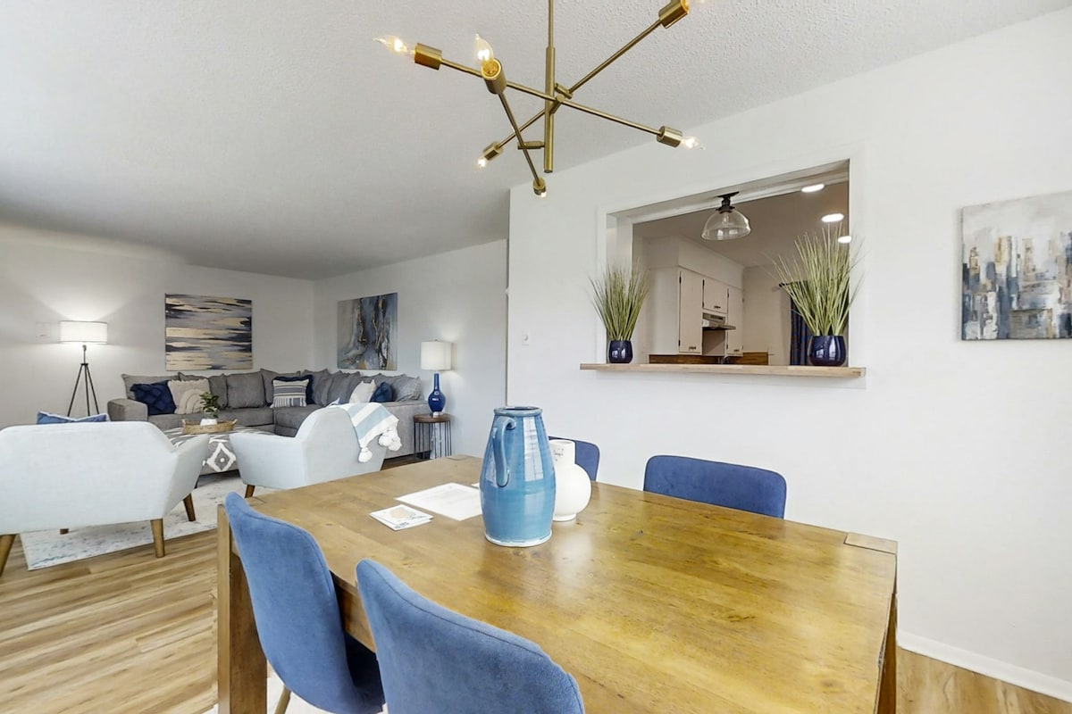 A dining area features a wooden table surrounded by blue upholstered chairs. A blue vase is positioned at the center of the table. In the background, a cozy living space includes a large sectional sofa and decorative wall art, creating a welcoming atmosphere.