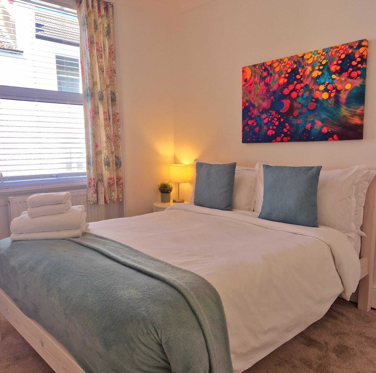 A cozy double room is highlighted by a spacious bed dressed in crisp white linens and soft blue accents. A colorful piece of wall art adds a vibrant touch above the bed. Soft natural light filters through a window adorned with floral curtains, illuminating the inviting space.