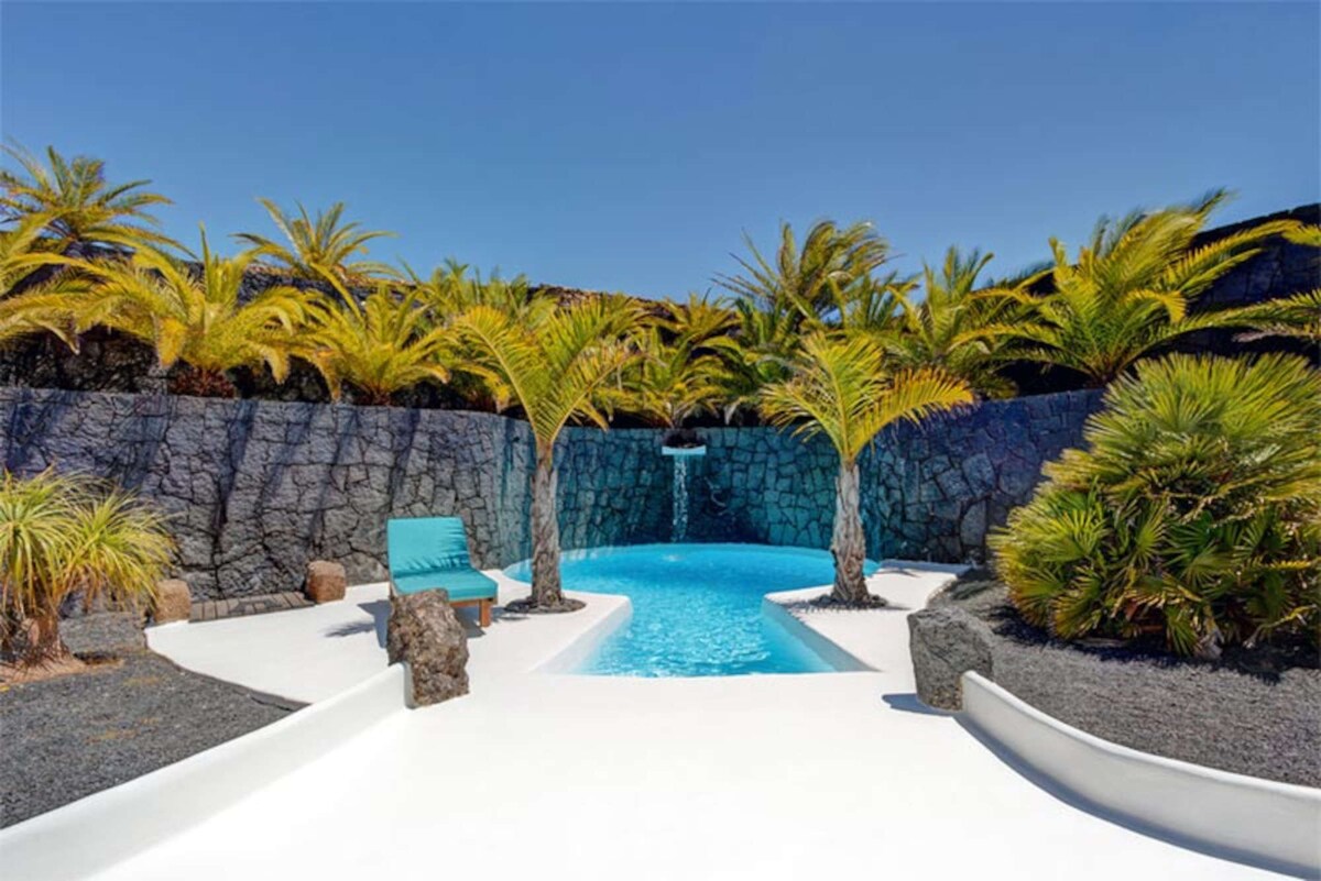 A serene outdoor space is presented, featuring a swimming pool surrounded by tropical palm trees. A stone wall encircles the area, with contrasting dark volcanic rock and white pathways leading to the pool. Sun loungers offer a place to relax in the warm sunlight.
