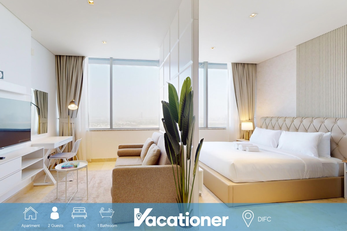 An open-plan living and sleeping area features a spacious queen-sized bed dressed in soft linens, a two-seater sofa for relaxation, and a smart TV mounted on the wall. Large windows provide ample natural light and offer city views, enhancing the modern ambiance.