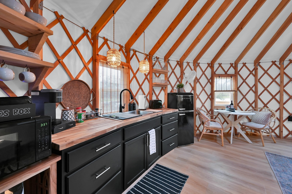 The kitchenette is designed with wooden cabinetry and a stylish countertop. It features a microwave, fridge, and cooking appliances. A dining table with chairs is positioned by a window, while woven lamps provide soft light, enhancing the cozy atmosphere.
