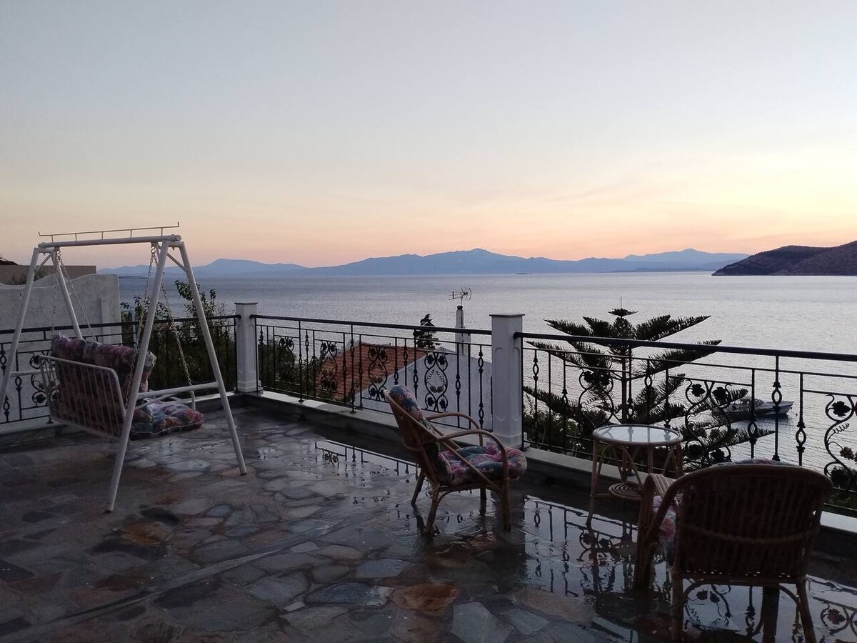 A spacious terrace offers a stunning view of the sea and distant mountains. Comfortable seating arrangements include a swing and two wicker chairs, surrounded by lush greenery and a stone floor reflecting the colors of the sunset.