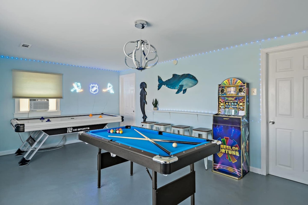 A game room features a pool table at the center, with an air hockey table against one wall. Neon artwork adds playful energy, while a vintage arcade machine stands nearby. A modern chandelier hangs overhead, and seating is available along the adjacent wall.