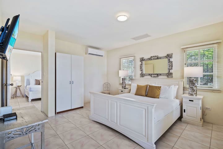 - חדר שינה מואר ואוורירי בגודל קווין סייז עם מראה מקושטת גדולה מעל המיטה
- אור טבעי זורם פנימה דרך החלונות, ומדגיש את העיצוב המינימליסטי
- דלת מובילה לחדר סמוך, ויוצרת סלון מחובר