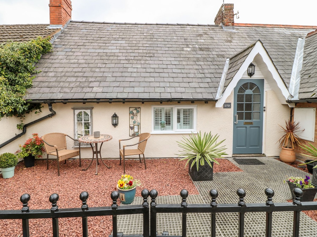 A charming front terrace features two wicker chairs and a small round table, surrounded by a gravel area with potted plants. The entrance is accentuated by a light blue door and decorative wall elements, providing an inviting first impression.