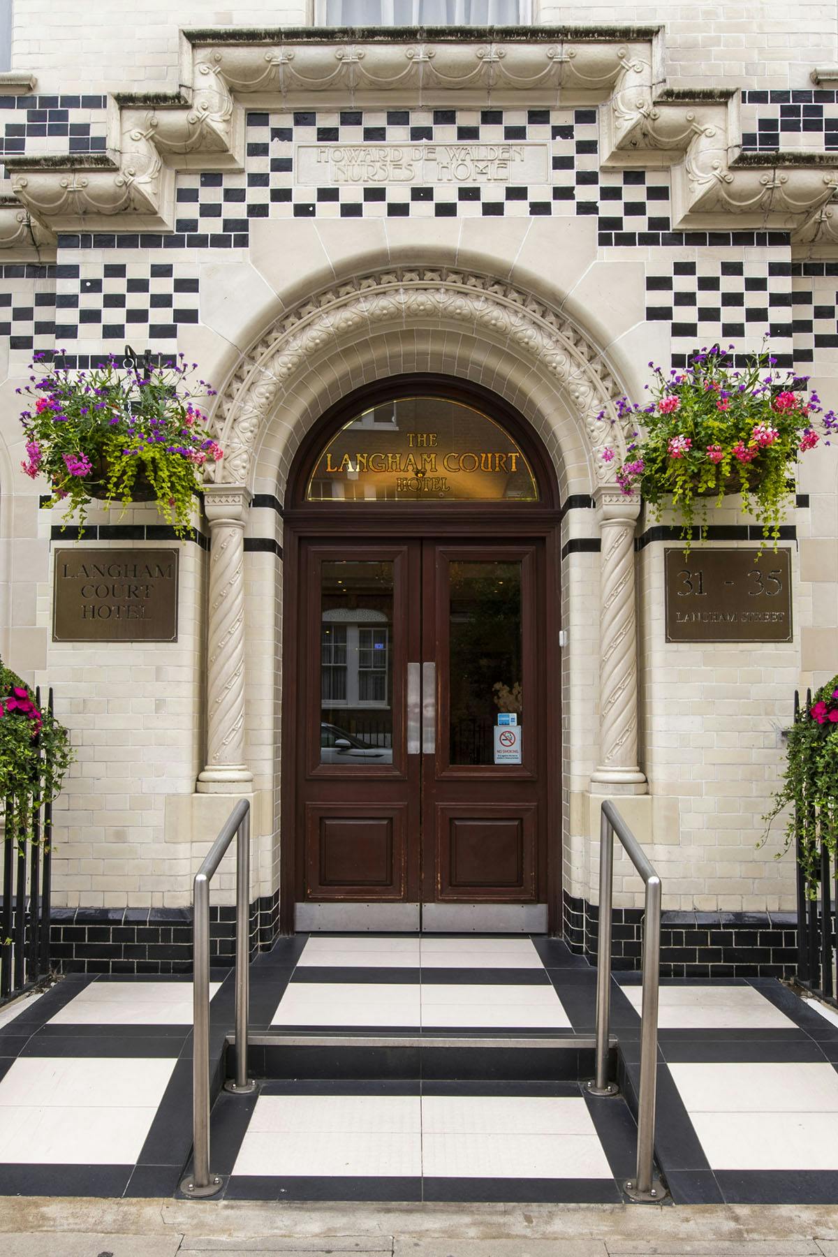 The entrance of the Langham Court Hotel showcases intricate architectural details, including decorative stonework and bold black and white tiles. Lush flower baskets hang beside the door, welcoming guests to the space with a blend of modern elegance and Victorian charm.