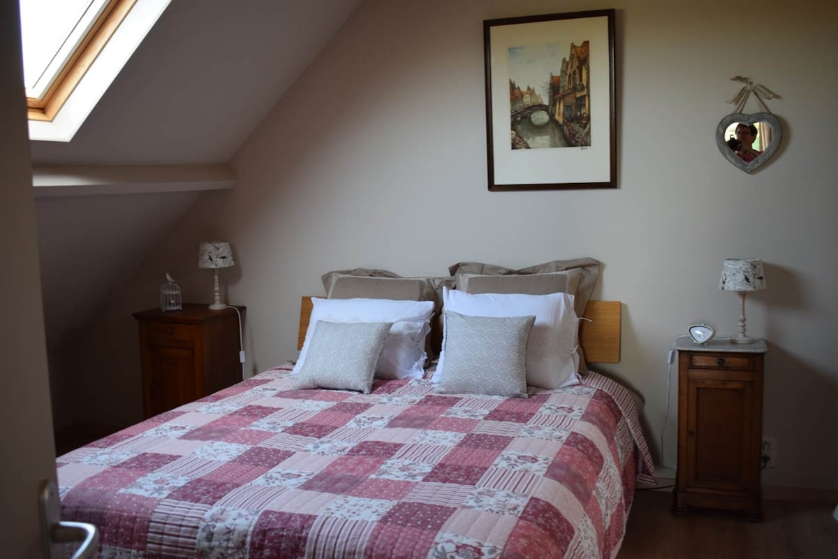 The sleeping area features a large bed dressed in a patterned quilt, complemented by two decorative pillows. Artwork hangs above the bed, and bedside tables are positioned on either side, each with a lamp. Natural light enters through a skylight.
