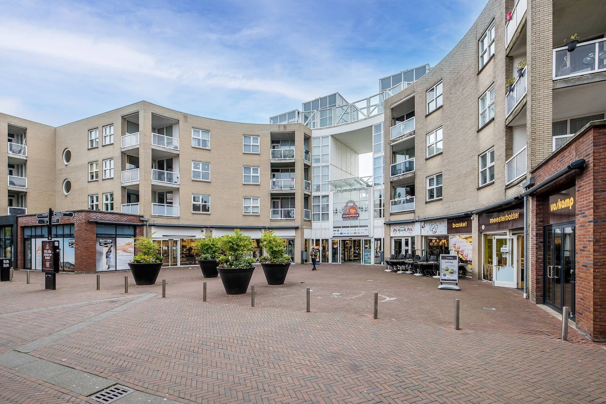 A modern plaza is depicted, featuring several multi-story buildings with large windows. Potted plants add greenery to the space. Various shops line the ground level, with outdoor seating visible, creating an inviting community atmosphere. The brick-paved area offers a clean and accessible layout.