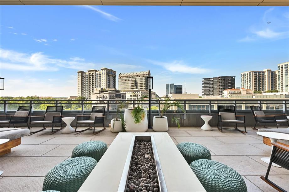 An expansive rooftop terrace is showcased, featuring modern seating options and green accent pillows. A linear planter filled with smooth stones runs through the center. Cityscape views are visible in the background, complemented by clear blue skies.