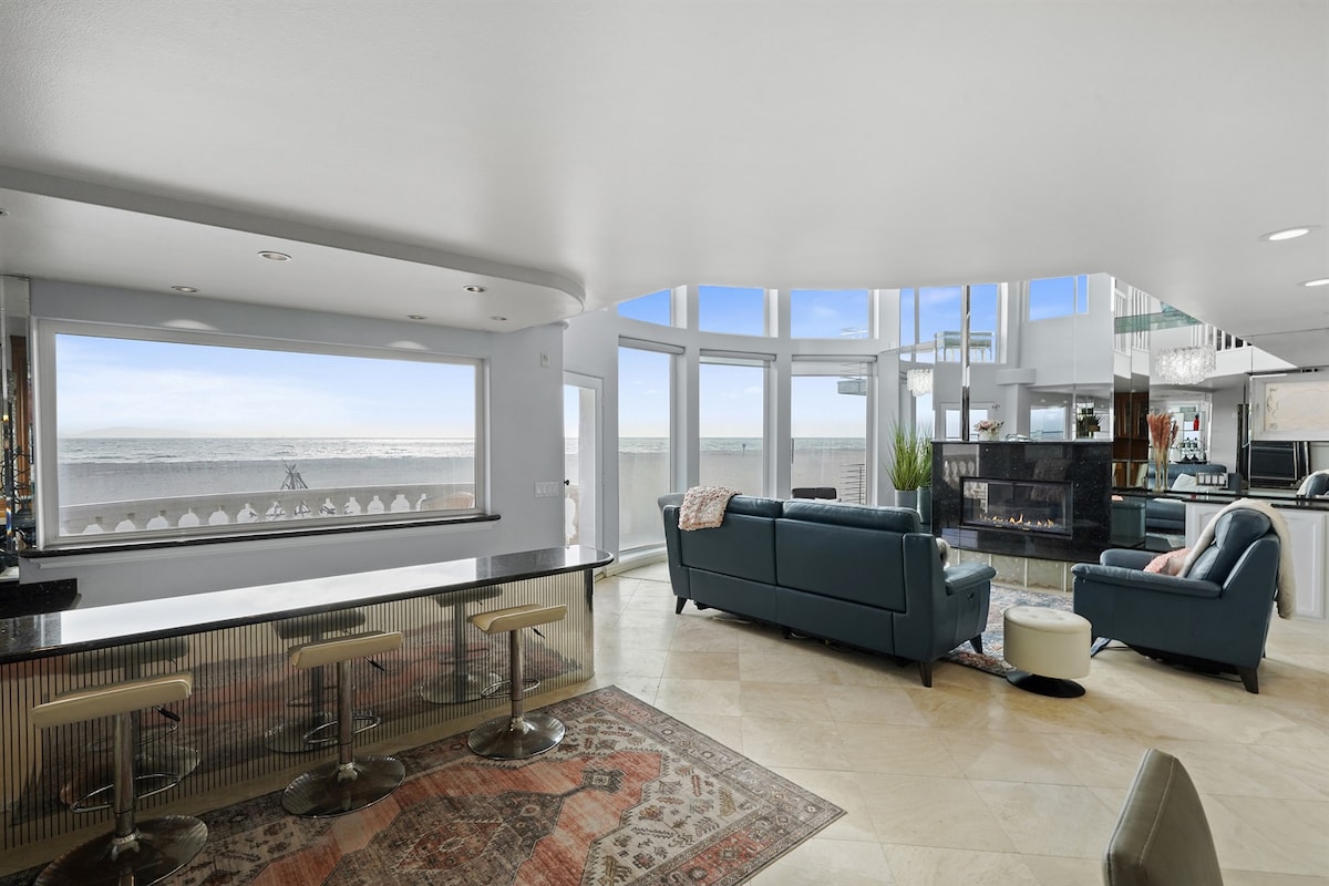 A spacious living area features large windows providing natural light and ocean views. Comfortable seating includes a sectional sofa, while a bar area complements the room. Tile flooring and a decorative rug enhance the ambiance, creating a welcoming coastal environment.