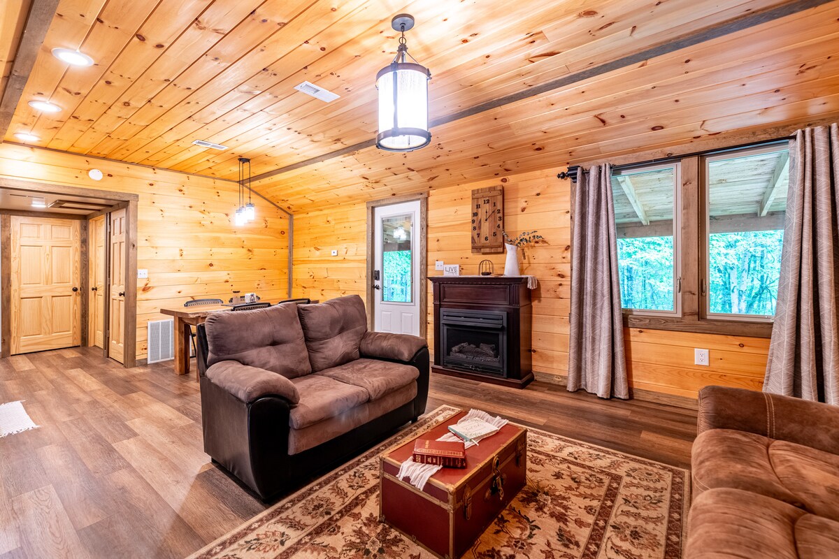 The open living area features natural wood-paneled walls and a cozy sofa, accompanied by a coffee table. A fireplace is situated against one wall, contributing to the warm ambiance. Large windows provide views of the outdoor surroundings, with curtains framing the daylight.