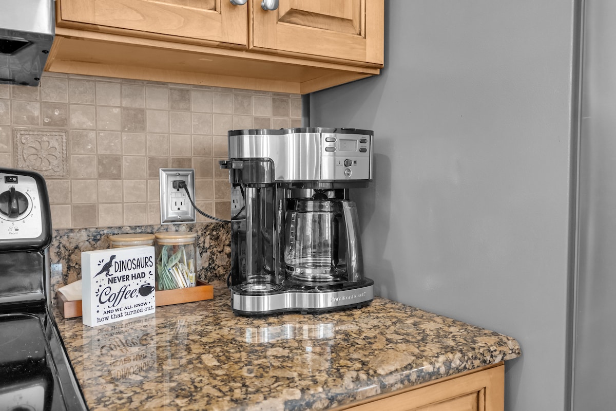 A modern coffee maker is positioned on a polished granite countertop, surrounded by warm wooden cabinetry. Soft lighting highlights the appliance and its glass carafe, enhancing the efficiency of the well-equipped kitchen where it is located.