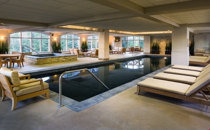 An indoor heated pool area features a dark water surface surrounded by wooden lounge chairs and a stone wall. Large windows provide natural light, illuminating the space and the landscaped greenery outside. Additional seating is arranged nearby, creating a relaxing environment.