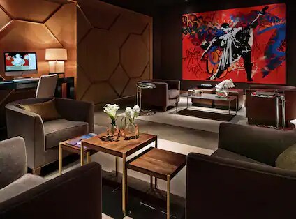 A modern seating area is furnished with plush chairs and stylish side tables topped with decorative vases. Bold artwork adorns the wall, and ambient lighting creates a relaxed atmosphere. A sleek desk with an electronic device is visible in the background.