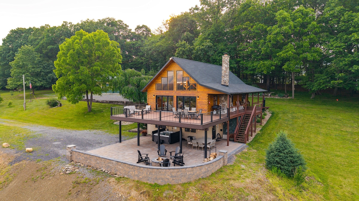 A spacious two-story retreat is set amidst a lush landscape, featuring a large balcony and multiple outdoor seating areas. The natural wood finish of the exterior complements the surrounding greenery, creating a serene atmosphere for relaxation.