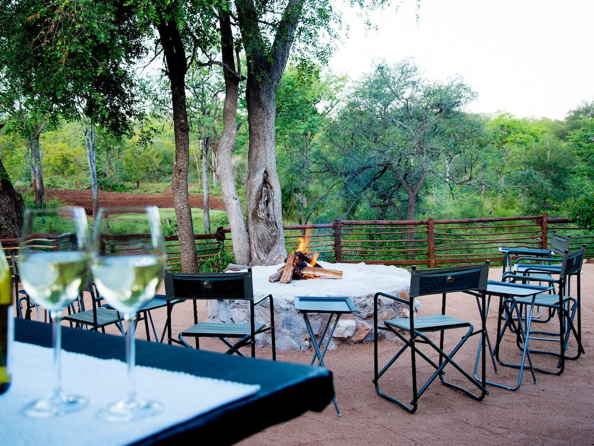 A communal fire pit is surrounded by several folding chairs, set on a sandy surface. The warm glow of the fire illuminates the area. Nearby tables hold drinks, complementing the natural surroundings of trees and greenery.