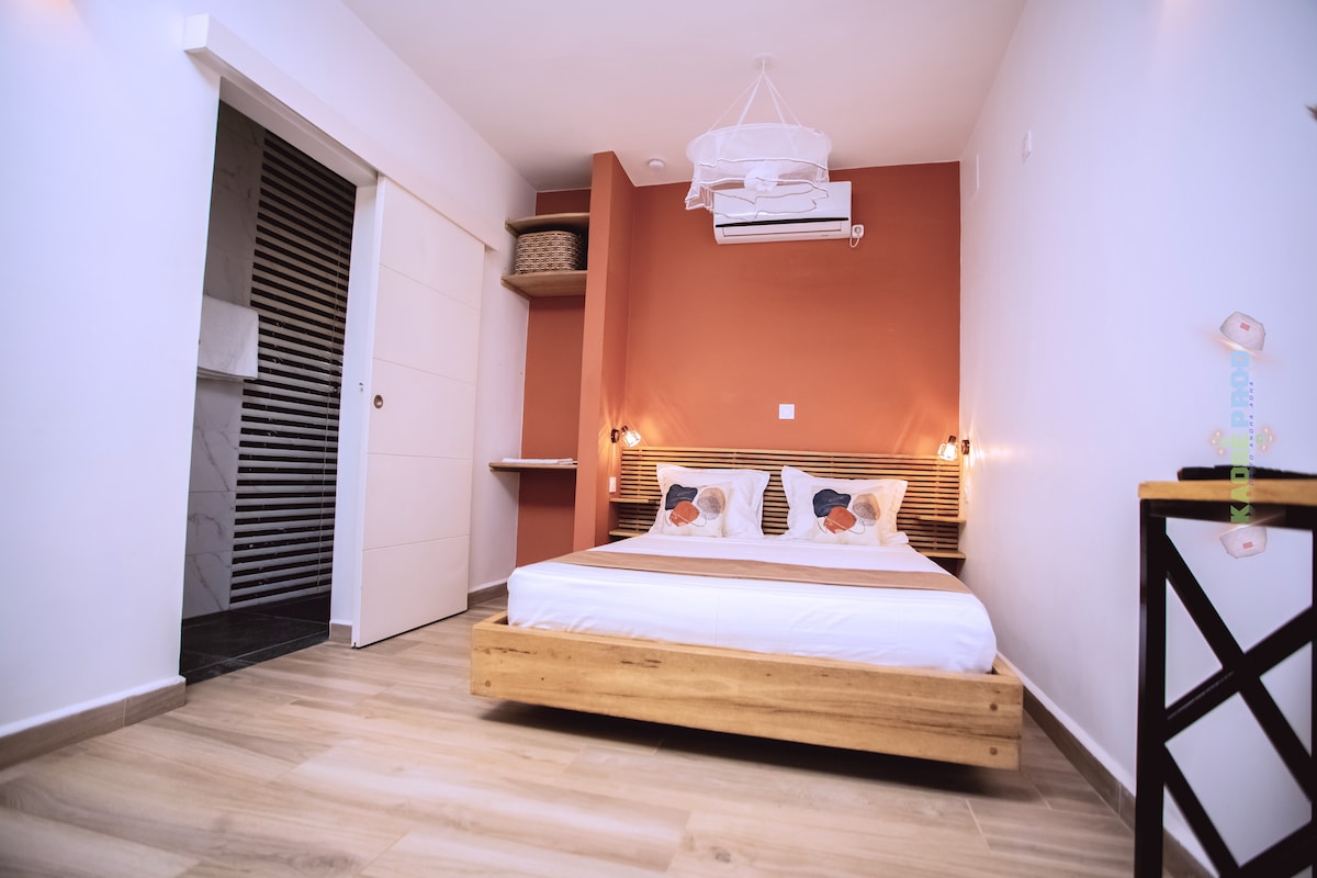 A well-appointed sleeping area features a spacious bed with neutral bedding, accented by colorful pillows. Soft lighting from wall sconces complements the warm terracotta wall behind. Natural wood elements and a small shelf add practicality, while a door to the left leads to additional space.