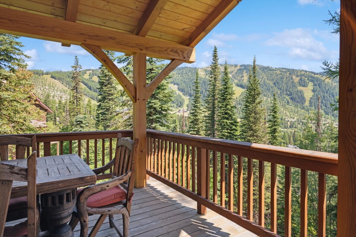 An outdoor deck is highlighted by rustic wooden beams and railings. A dining table with matching chairs occupies the space, offering views of the surrounding forested mountains and ski slopes, framed by tall evergreen trees under a clear blue sky.