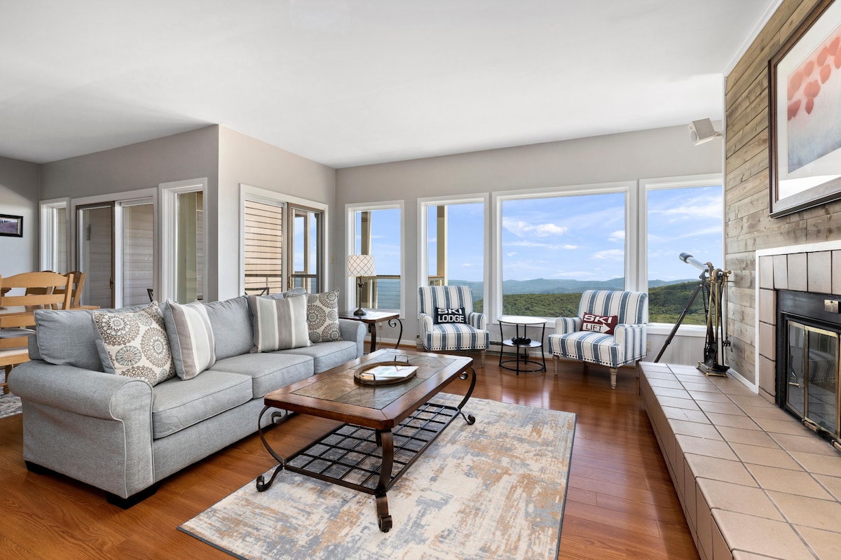 A spacious living area features a large sectional sofa adorned with patterned cushions, complimented by a coffee table centered on a decorative area rug. Large windows provide scenic views of the surrounding landscape, and a fireplace adds an element of warmth.