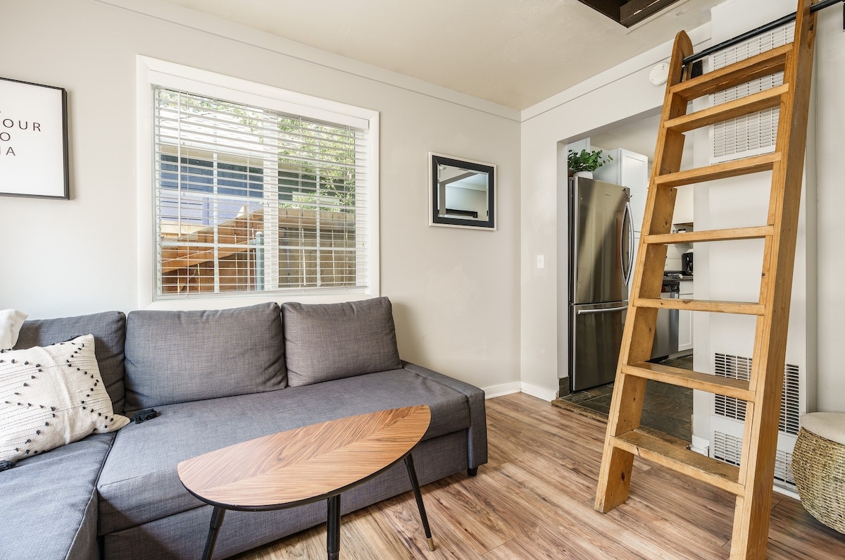 A cozy living area features a grey couch with a chaise lounge and a modern wooden coffee table. A wooden ladder leads to the upstairs loft, while natural light streams through large windows. The space is adorned with simple decor and a framed mirror.