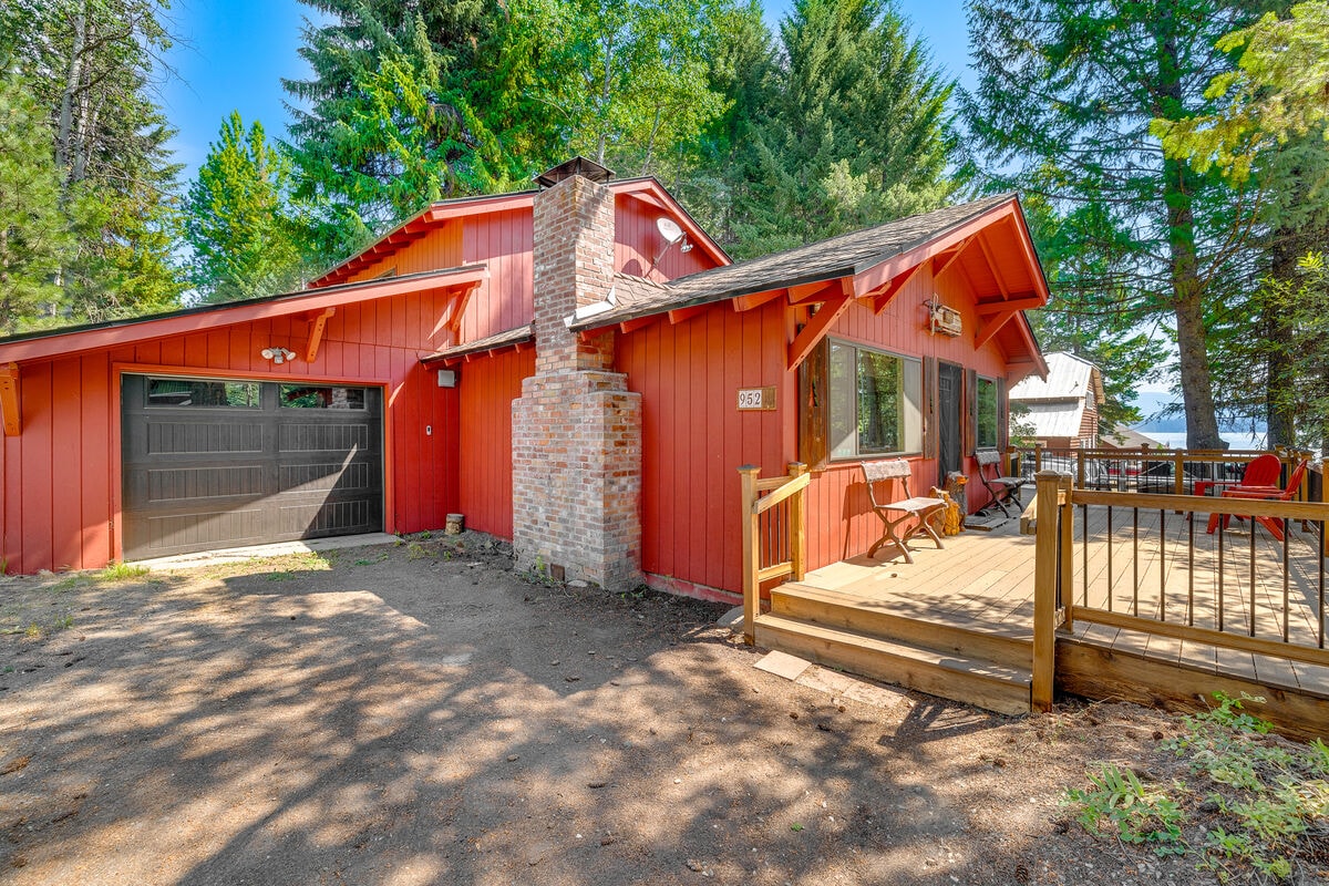 A charming cottage is shown with a vibrant red exterior, surrounded by tall green trees. A wooden deck provides access to outdoor seating, and a brick chimney adds character. The image captures the inviting entrance and the garage area, with sunlight filtering through the foliage.