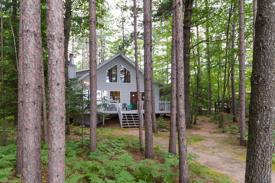 A charming lakefront retreat is partially visible among tall trees, with a light grey exterior and a welcoming porch. The path leading toward the house is flanked by lush greenery, enhancing the serene natural setting.