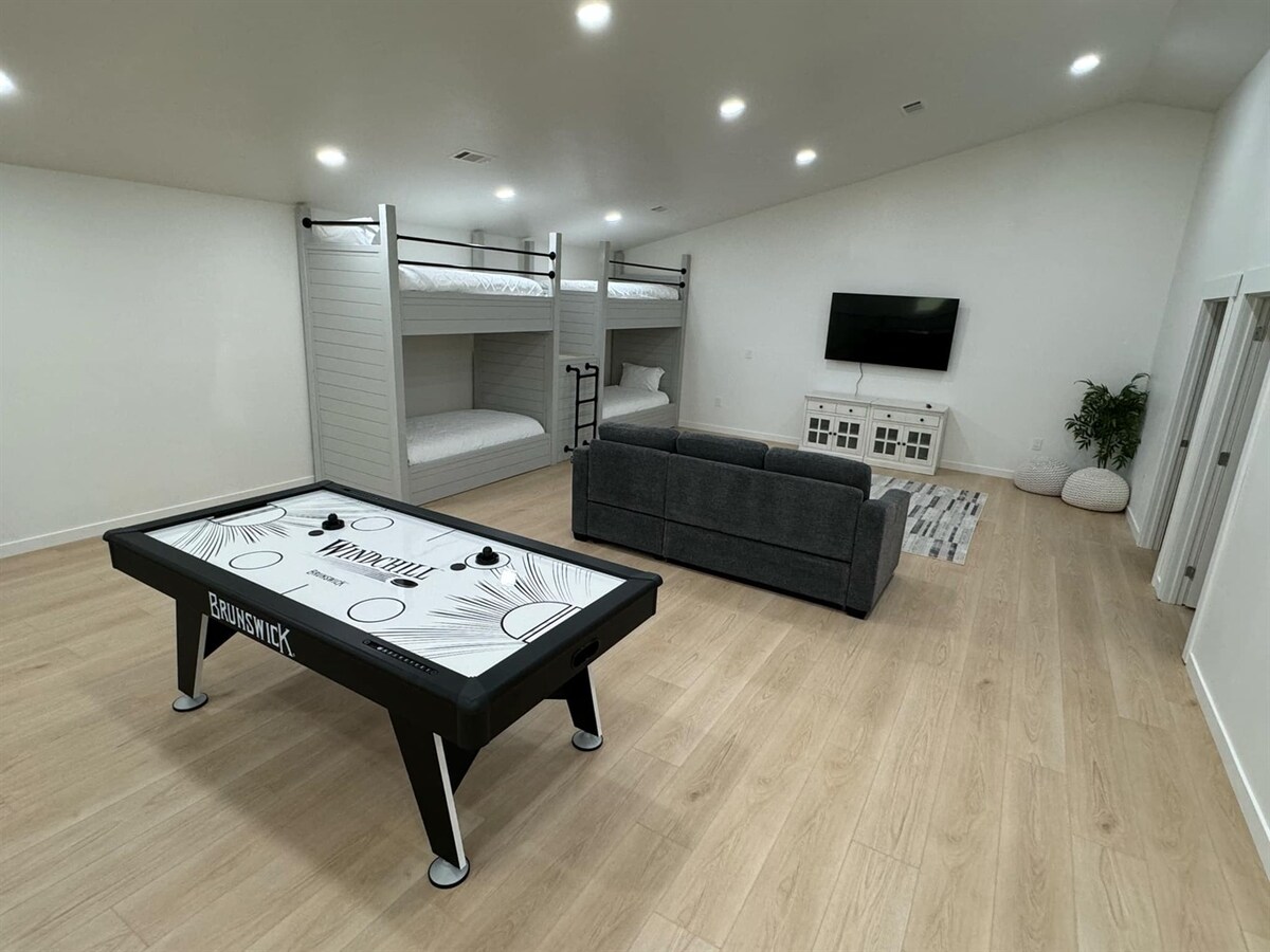 An indoor recreational area features a sleek air hockey table at the center, surrounded by light wood flooring. A cozy sectional sofa is positioned nearby, with a large wall-mounted television and white cabinetry in the background. Two sets of bunk beds are located against the wall.