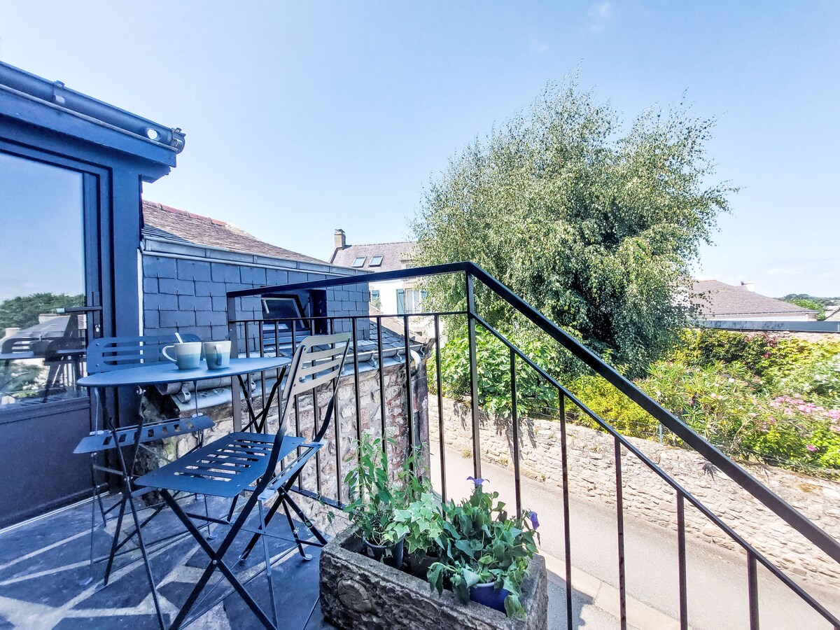 A balcony area features a small, round table with two metal chairs, ideal for outdoor relaxation. A planter with greenery adds a natural touch. The view includes nearby trees and a peaceful street, contributing to a serene environment.