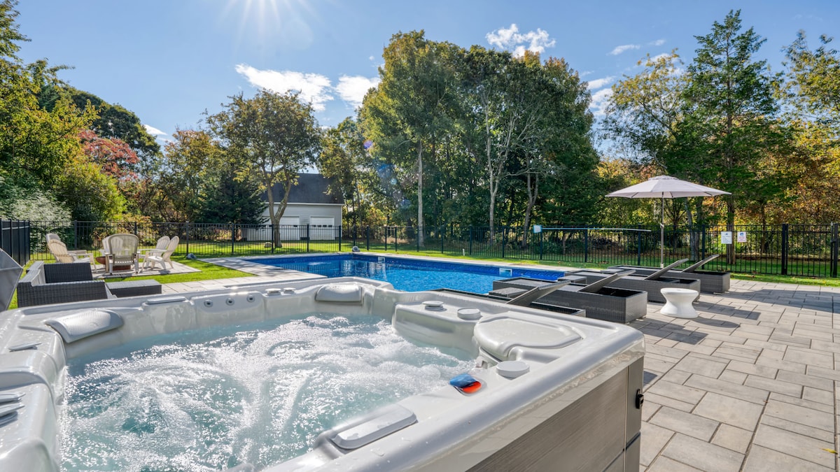 A freestanding hot tub is positioned beside a private heated pool, surrounded by a spacious patio area. Lounge chairs and a parasol are positioned nearby, with greenery providing a natural backdrop. The sunlight creates a bright and inviting atmosphere.