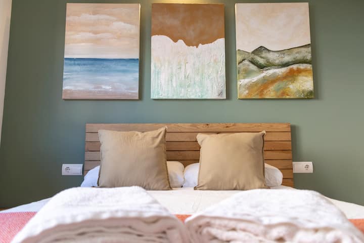 Ovaj pogled izbliza na spavaću sobu usredotočen je na pažljivo osmišljene detalje koji ovaj smještaj čine doista privlačnim. Drveno uzglavlje s rešetkama daje dašak topline i prirodne teksture, savršeno nadopunjujući trio mirnih pejzažnih umjetničkih djela iznad.