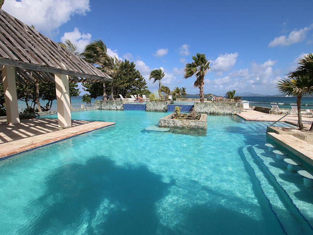 A refreshing pool area is surrounded by swaying palm trees and offers views of the ocean beyond. The water glistens under a sunny sky, and a shaded pavilion is visible on one side, providing a relaxing place to unwind.