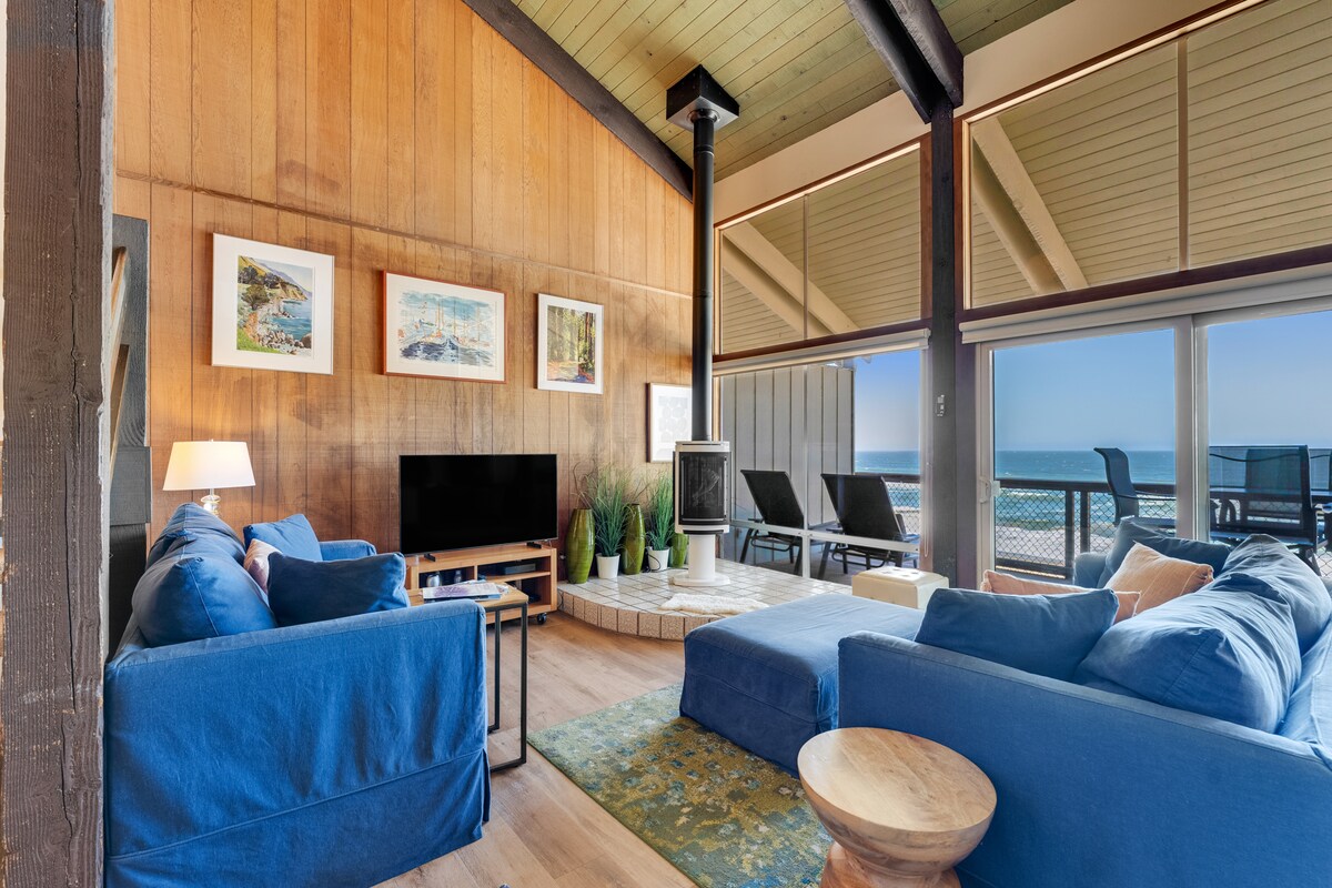 A bright living area features two blue sofas arranged around a light wood coffee table. Wall art is displayed above a television, and a gas fireplace is positioned nearby. Large windows provide coastal views, while natural light fills the space through the elevated ceiling.