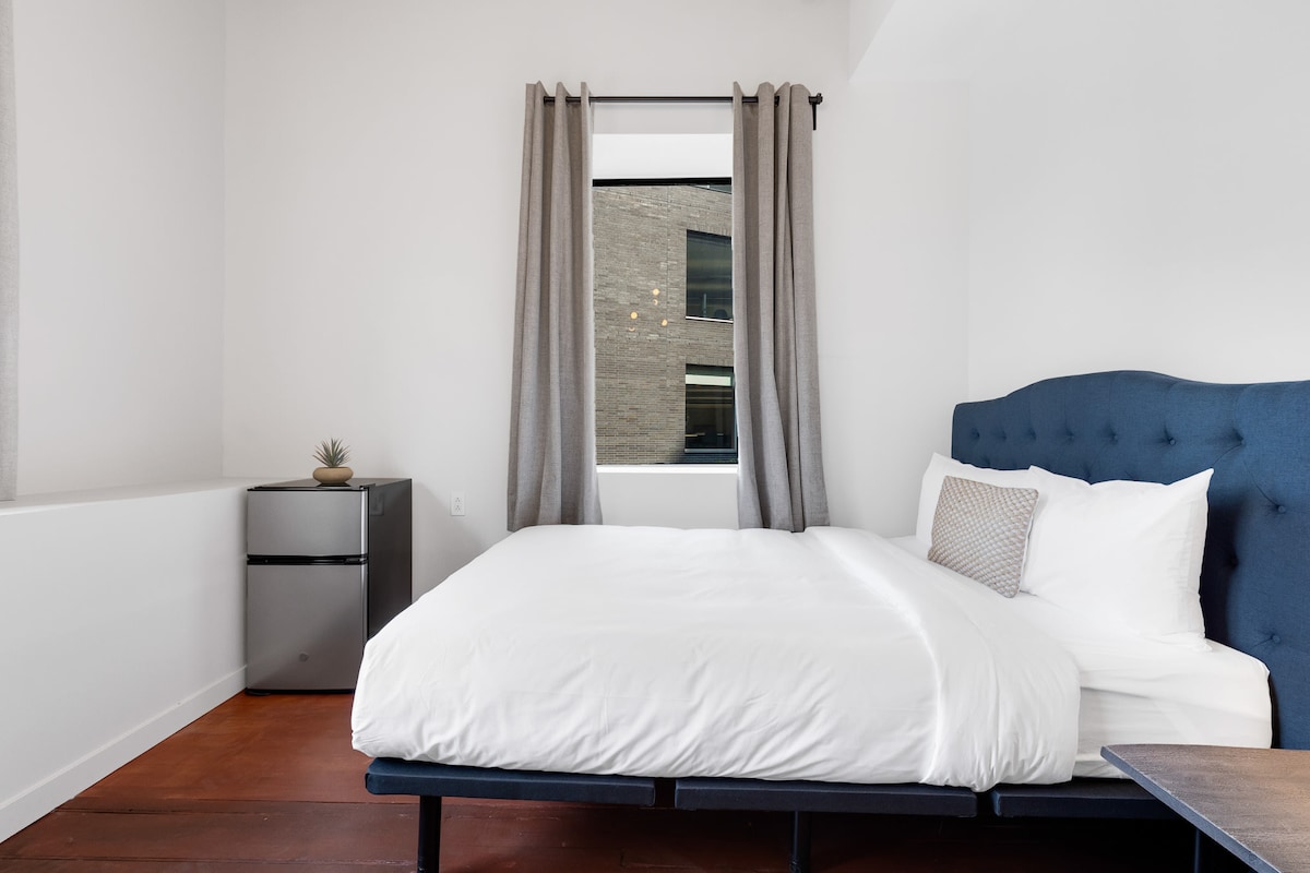 A bright guest room features a spacious bed with crisp white linens and a decorative pillow. Soft gray curtains frame a large window, allowing natural light to fill the space. A small refrigerator is positioned beside the bed, contributing to the room's functionality.