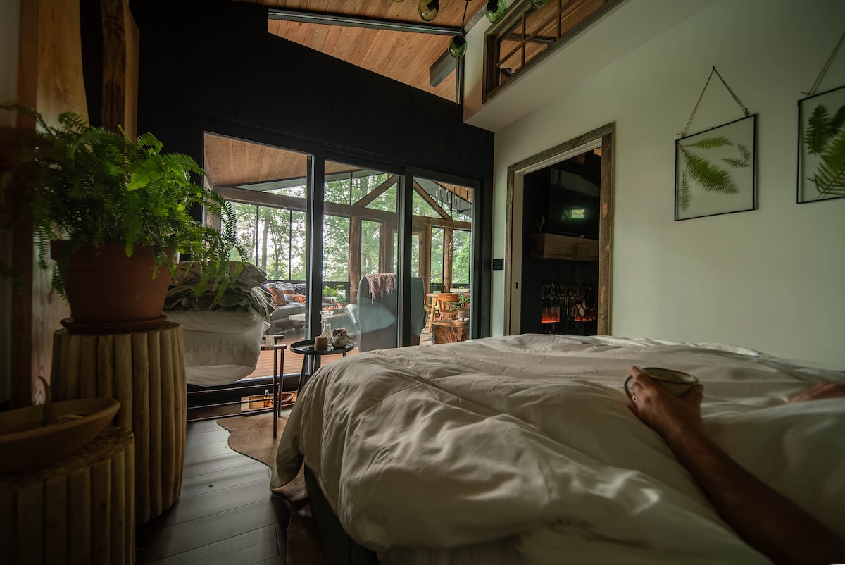 The inviting master bedroom features a spacious king-size bed draped in soft linens, with natural wood accents enhancing the decor. Large windows offer views of the serene screened-in porch, surrounded by lush greenery, creating a calming atmosphere. A cozy setting is brought to life by warm lighting.
