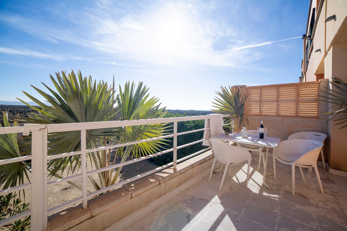 An outdoor terrace is displayed, featuring a round table set with white chairs. Lush green plants are visible, and the expansive view of the landscape is complemented by clear blue skies. Bright sunlight illuminates the space, enhancing the overall openness.