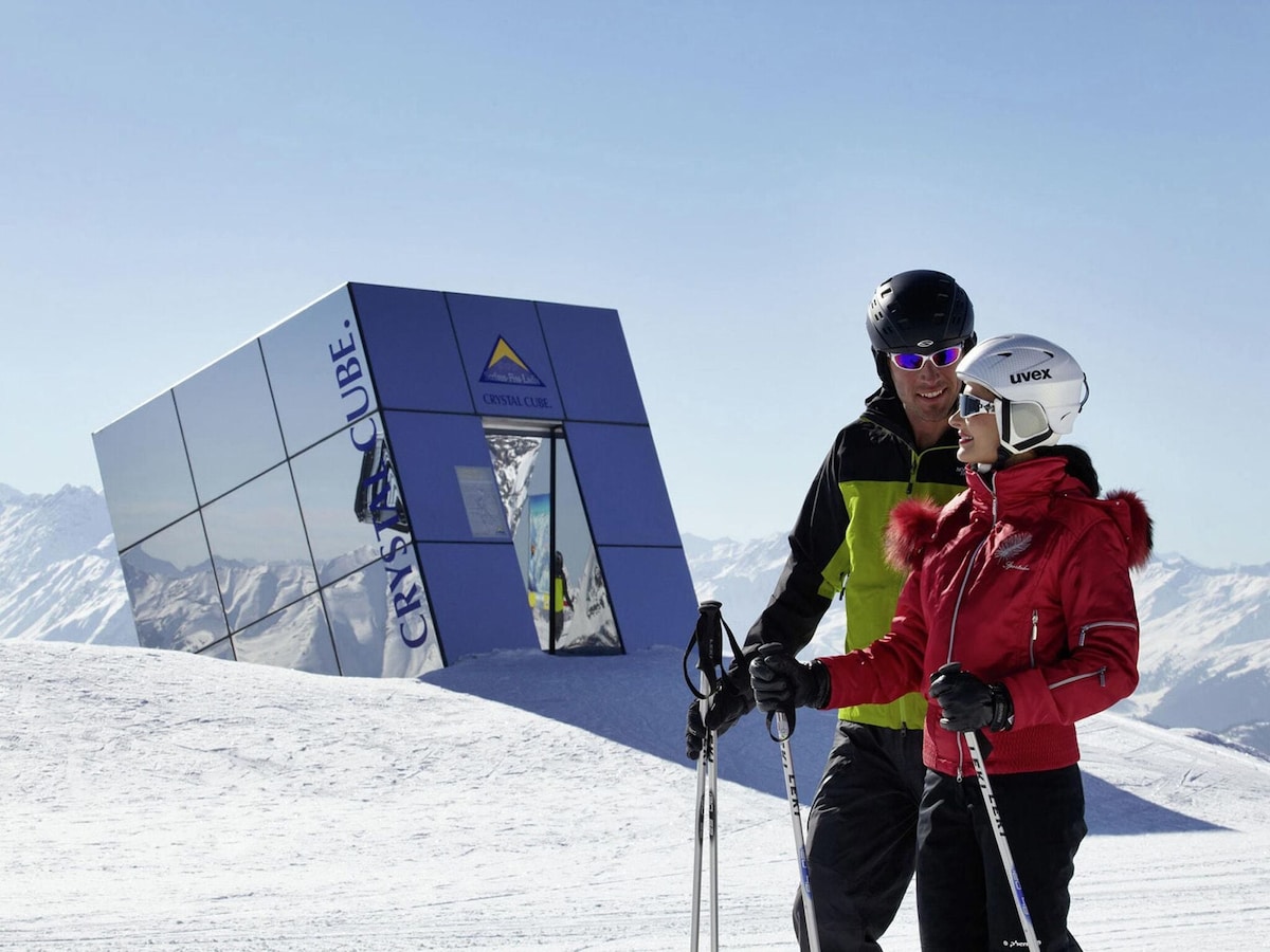 A modern, mirrored structure stands in a snowy landscape, reflecting the surrounding mountains. Two skiers dressed in colorful winter attire are positioned in the foreground, suggesting an active outdoor environment ideal for winter sports.