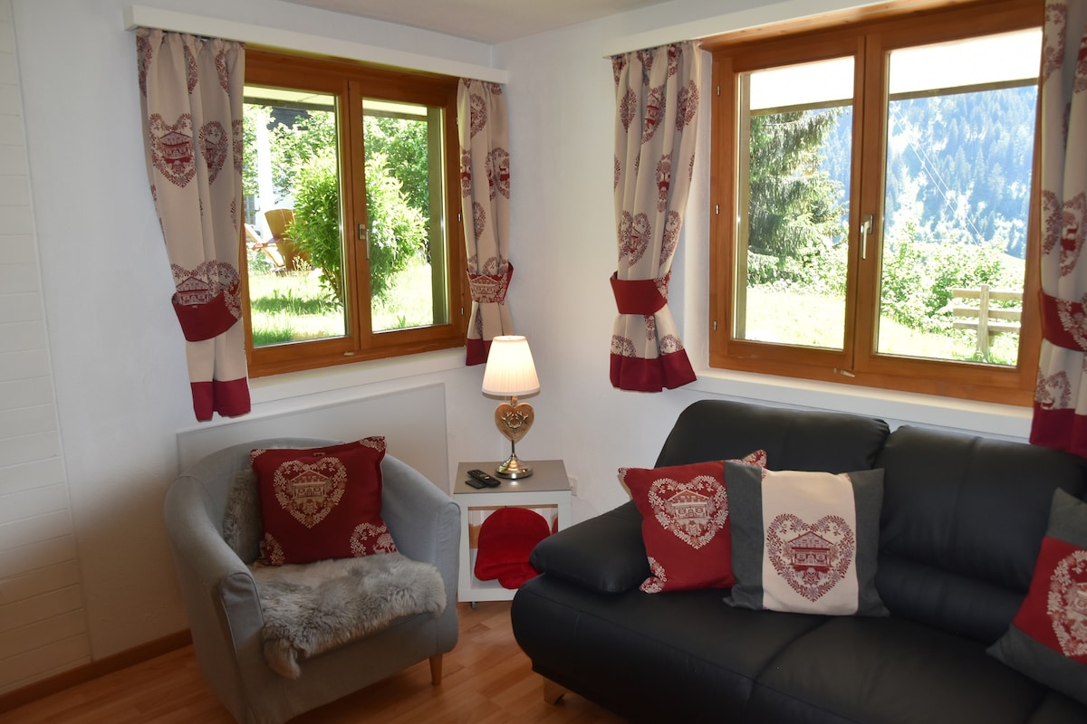 A comfortable living area is highlighted by two large windows offering views of the surrounding greenery. A dark sofa is accompanied by a cozy armchair and decorative pillows featuring heart designs. A small bedside table holds a lamp, adding to the inviting ambiance.