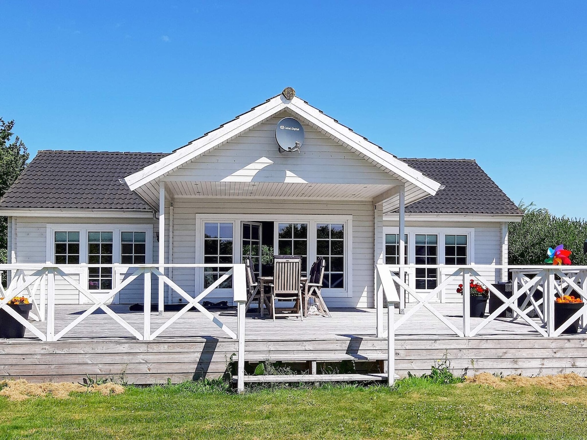 A bright holiday cottage features a spacious wooden terrace with a white railing, providing access to the indoors. The front is adorned with large windows, showcasing modern architecture and inviting outdoor seating. The surrounding natural area offers a serene backdrop.