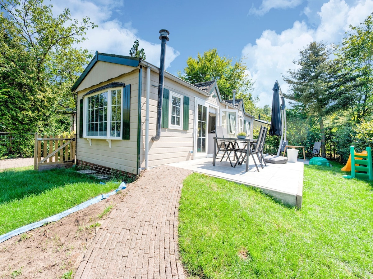 A charming chalet exterior is displayed, featuring large windows that invite natural light. A patio area with outdoor furniture is situated alongside a well-maintained lawn, framed by lush greenery and trees. A pathway made of bricks leads toward the entrance.