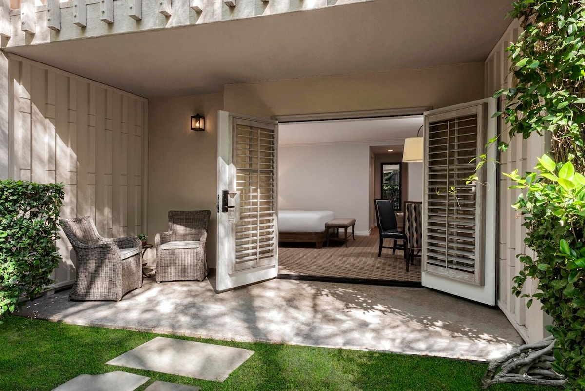 A private patio entrance is framed by lush greenery, featuring two comfortable chairs and a small table. Light spilling in through the open French doors reveals the spacious interior, where soft tones are complemented by natural light enhancing the tranquil ambiance.
