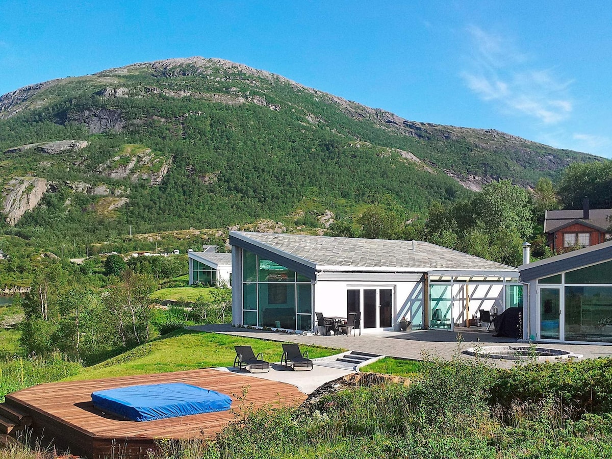 The holiday home is set against a scenic mountain backdrop, surrounded by lush greenery. A wooden terrace with lounge chairs is visible in the foreground, and the expansive glass windows of the house allow for a connection between indoor and outdoor spaces.
