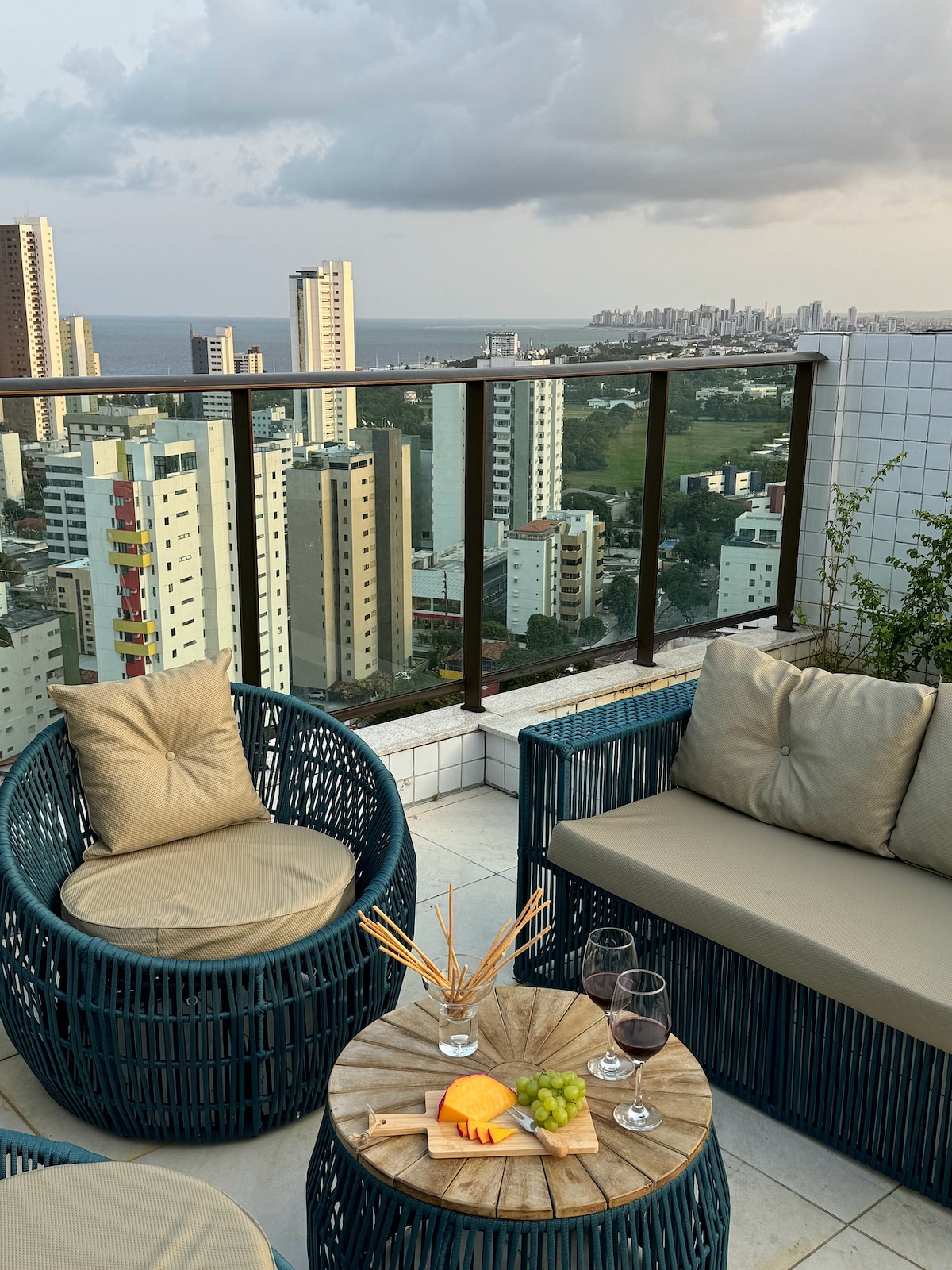 The rooftop space features cozy seating arranged around a wooden table. A selection of fruits and drinks is displayed, with a panoramic view of the city skyline and ocean in the background. The setting evokes a relaxed atmosphere, enhanced by surrounding greenery.